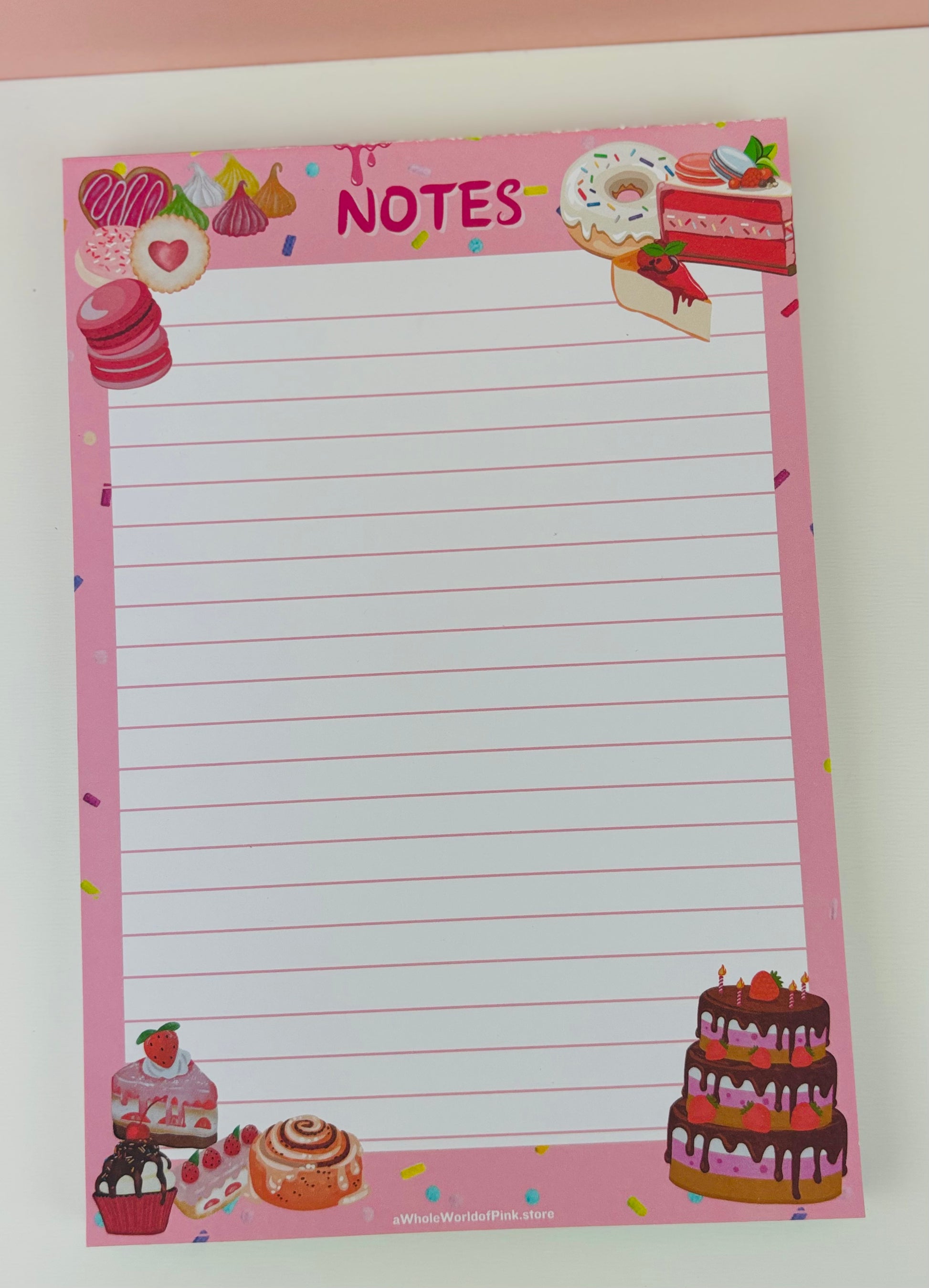 A5 Lined Note Pad Pink Patisserie Themed 50 Pages 120gsm, flat on white table with cakes, donuts, eclairs and sweets designed on pages, pov from above