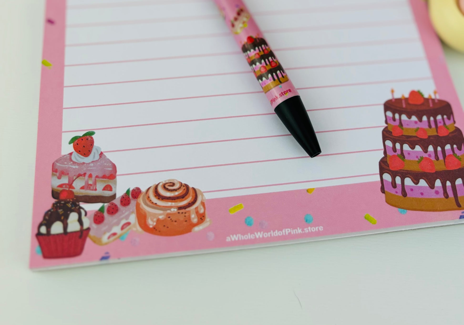 A5 Lined Note Pad Pink Patisserie Themed 50 Pages 120gsm, flat on white table with cakes, donuts, eclairs and sweets designed on pages, shot of footer of page with pen