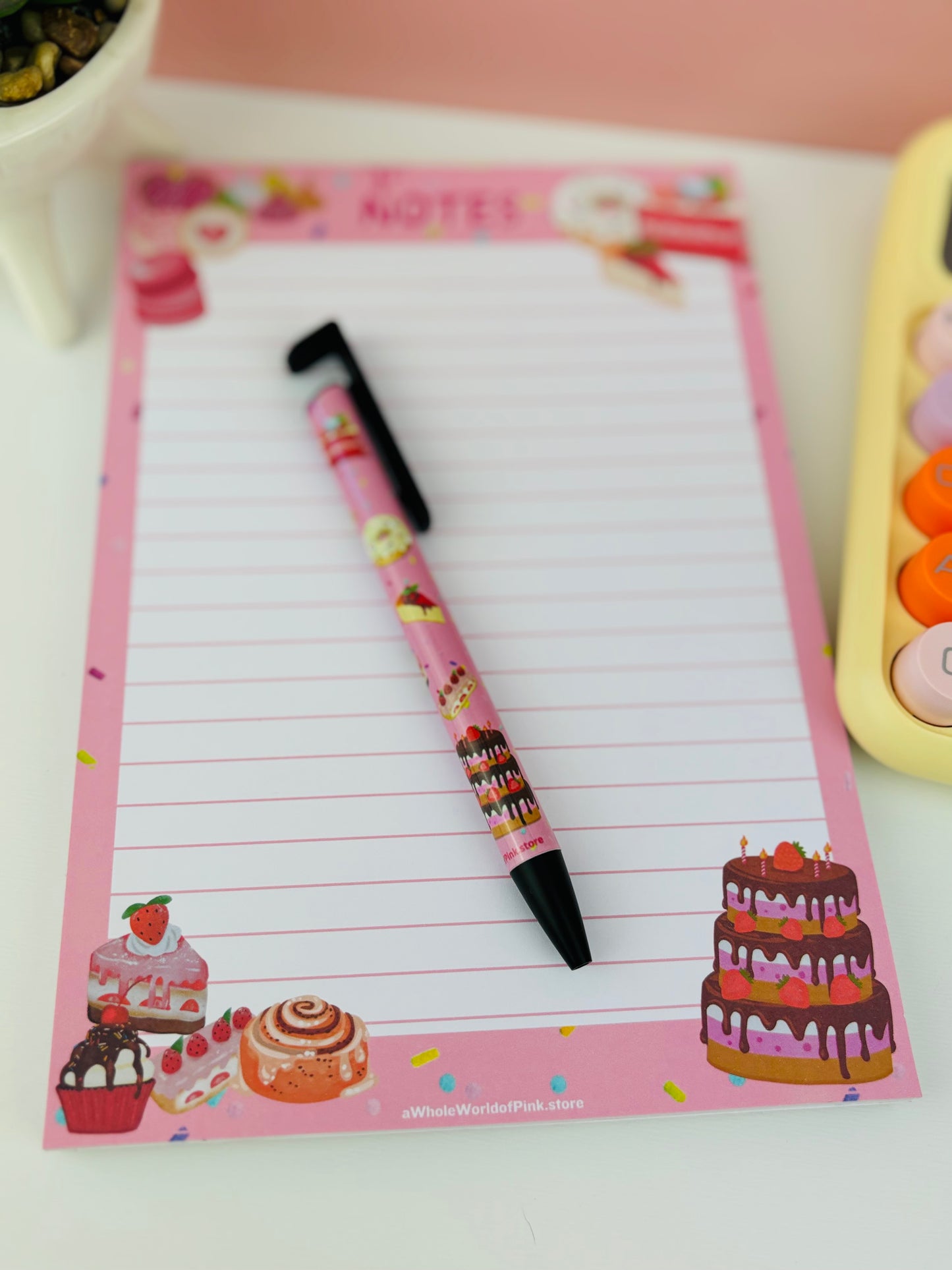 A5 Lined Note Pad Pink Patisserie Themed 50 Pages 120gsm, flat on white table with cakes, donuts, eclairs and sweets designed on pages, with pink patisserie themed pen and yellow calculator